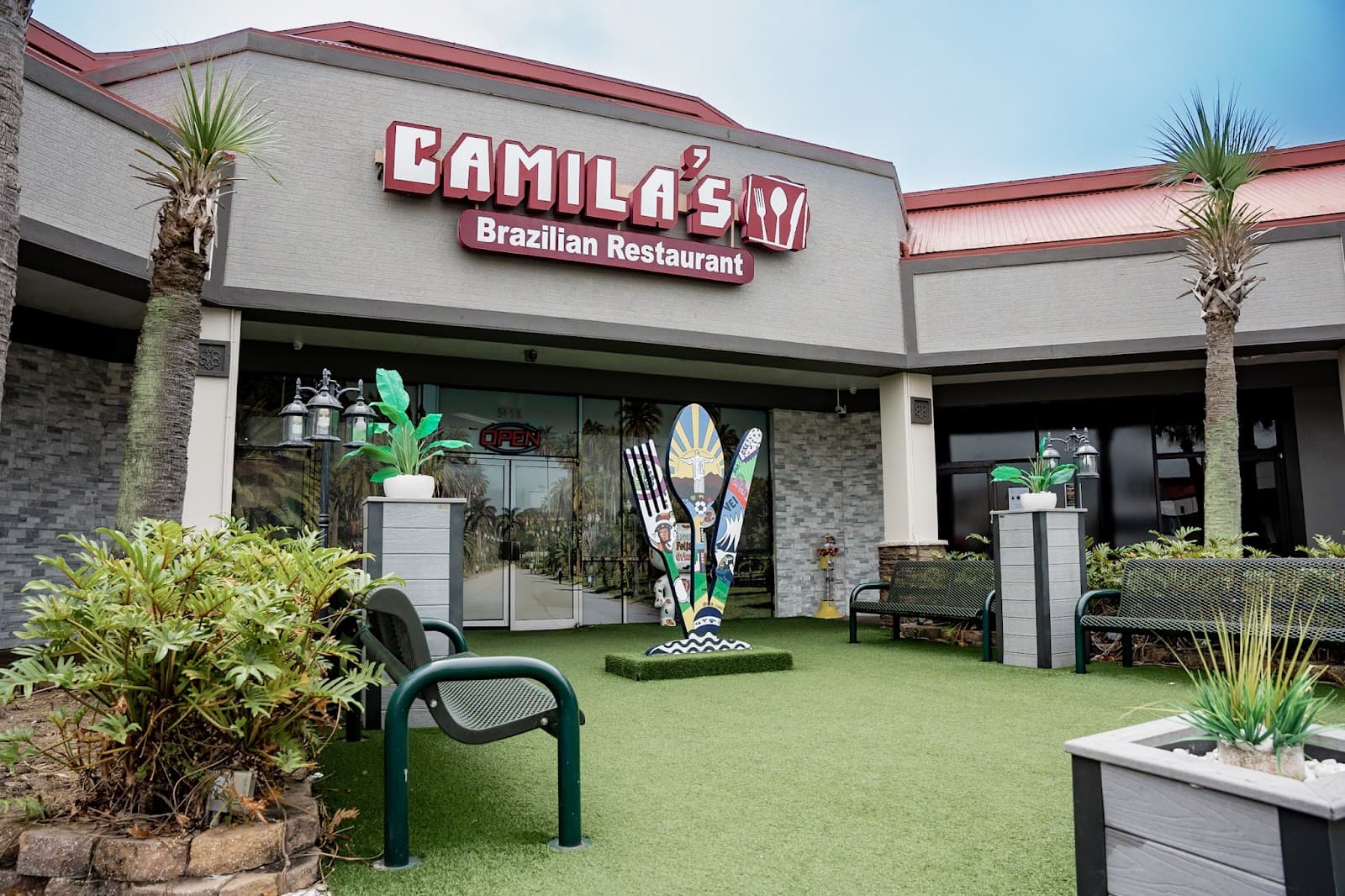 Fachada do Camila's na International Drive, com o letreiro vinho, o pátio de grama e o garfo e a colher gigantes na porta
