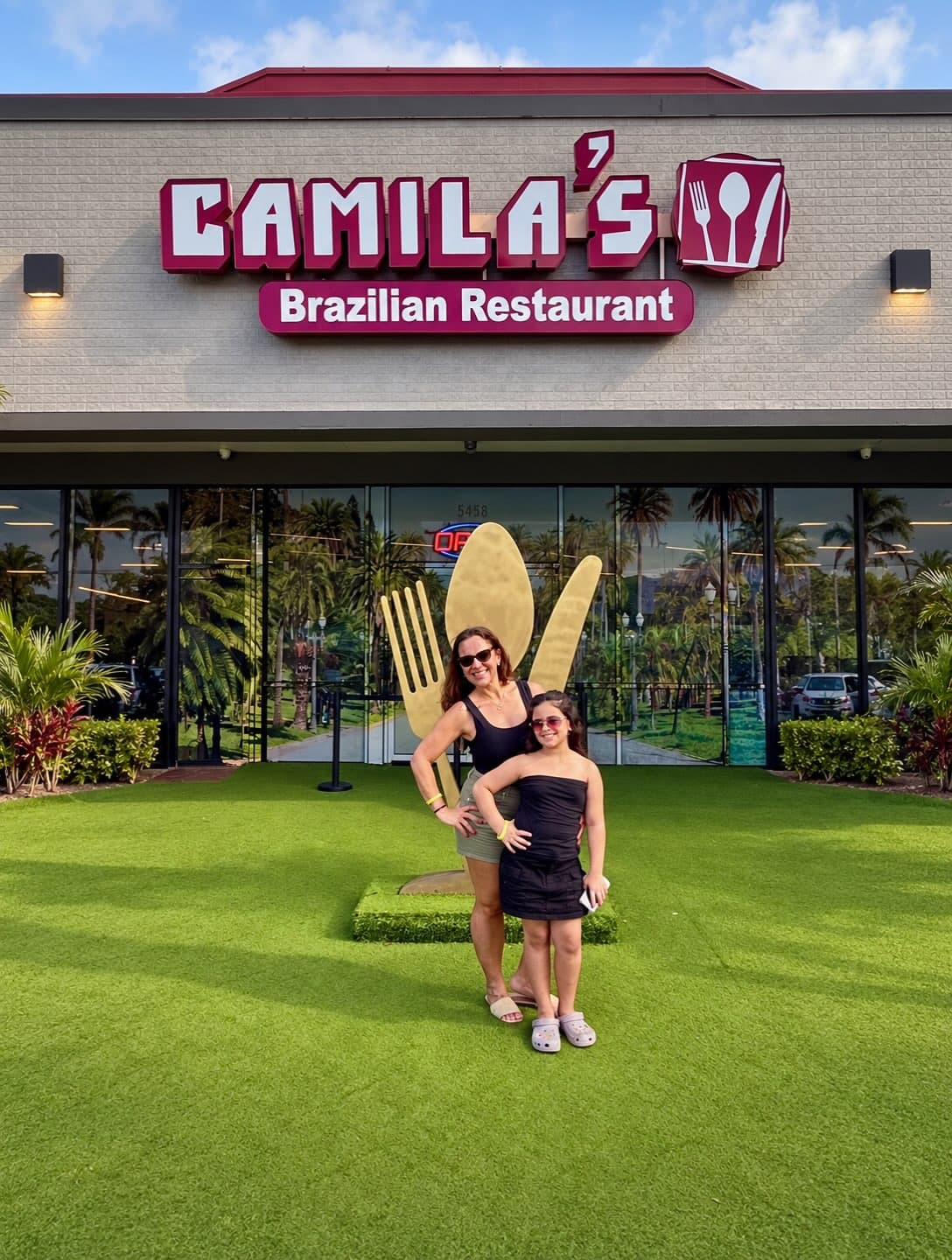 A entrada do Camila's: o letreiro, o número 5458 na porta, o garfo e a colher dourados e o gramado, com mãe e filha posando